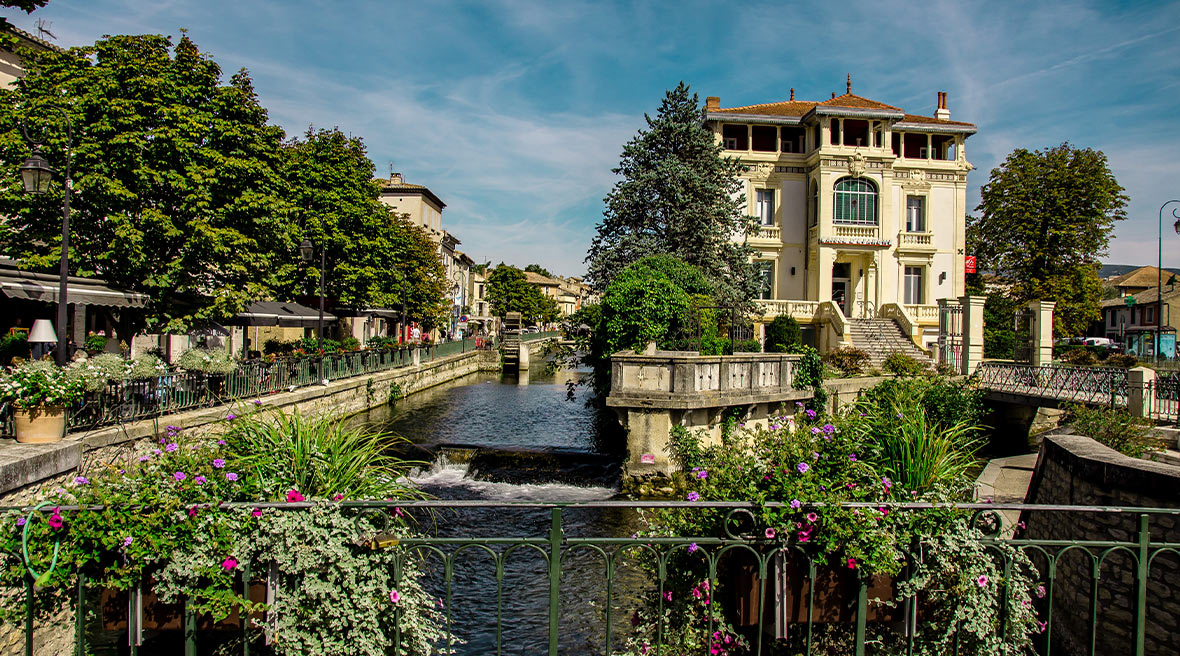 bridge overlooking a pretty French building with greenery and flowers surrounding a river flowing past